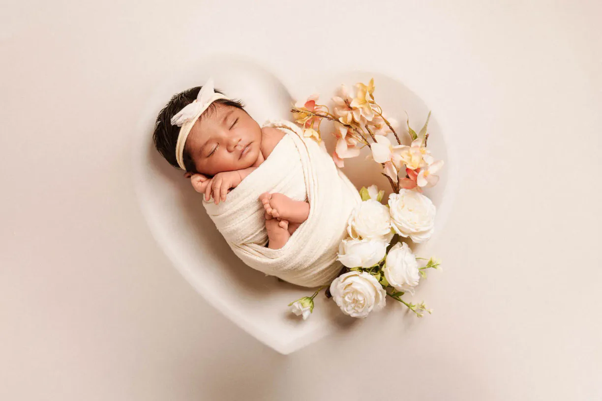 Newborn baby sleeping in a white heart-shaped bowl with flowers