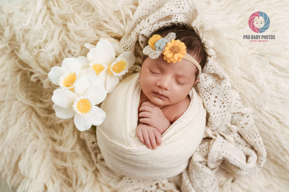 Newborn baby wrapped in white fabric beside large pale flowers